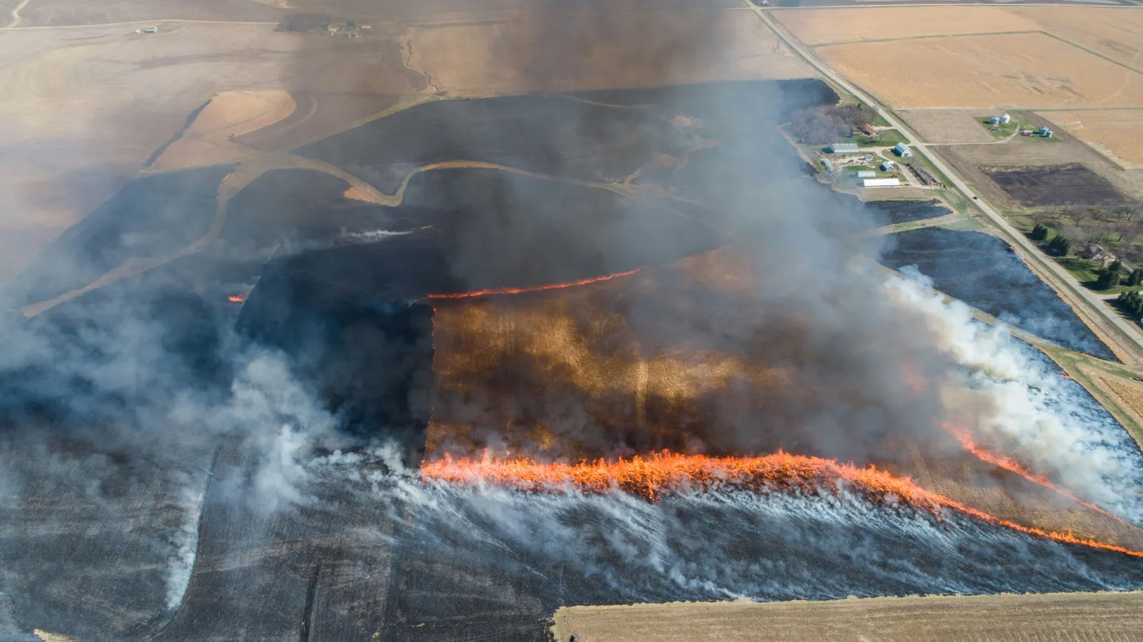 An aerial view of a bright fire line advancing across a geometric farm field, with smoke drifting over burned black ground and farmland beyond.